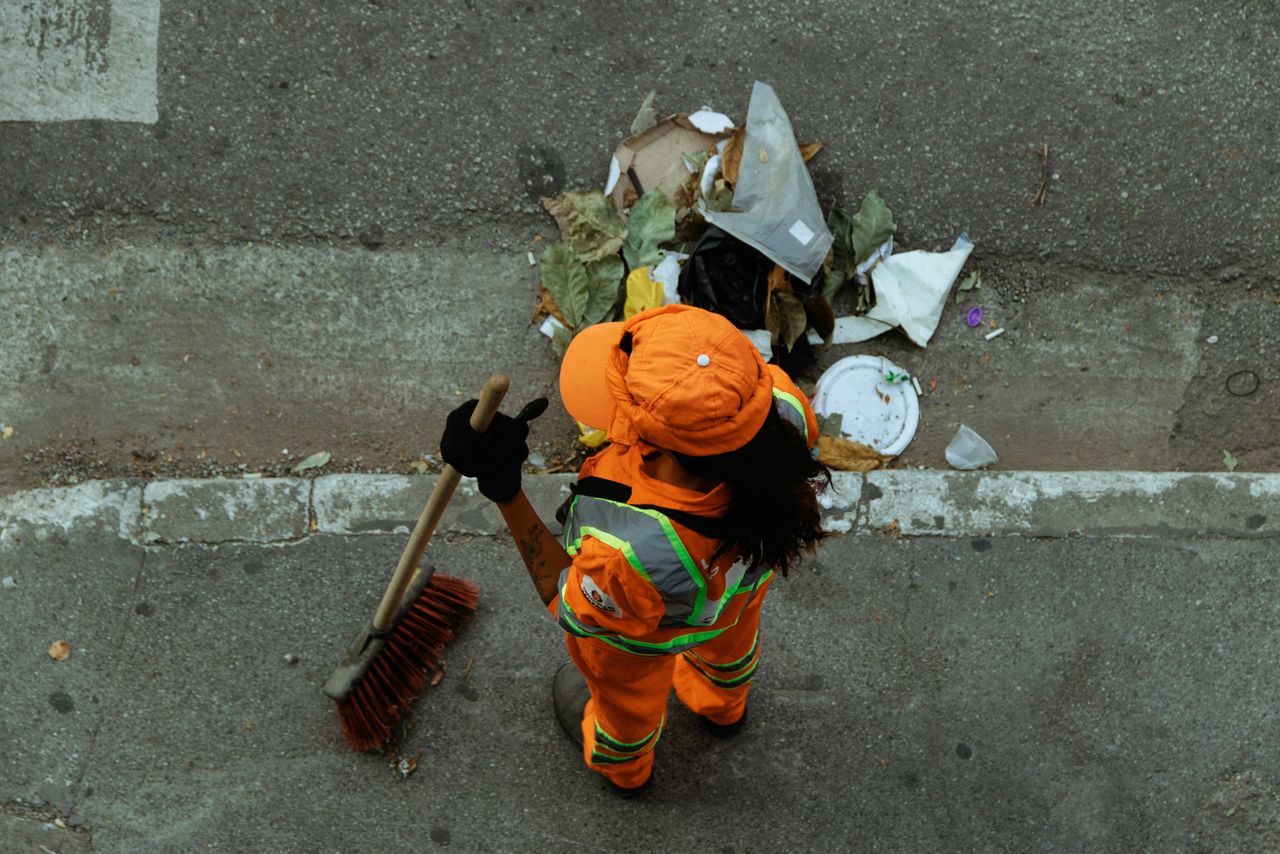 about-img Top view of a street cleaner wearing orange uniform sweeping debris on a city sidewalk.
